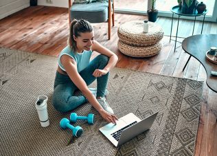 A sporty woman in sportswear is sitting on the floor with dumbbells and a protein shake or a bottle of water and is using a laptop at home in the living room. Sport and recreation concept.